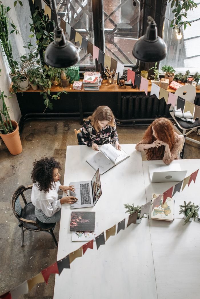 our-mission-01 Three women collaborate creatively in a cozy indoor workspace filled with plants.